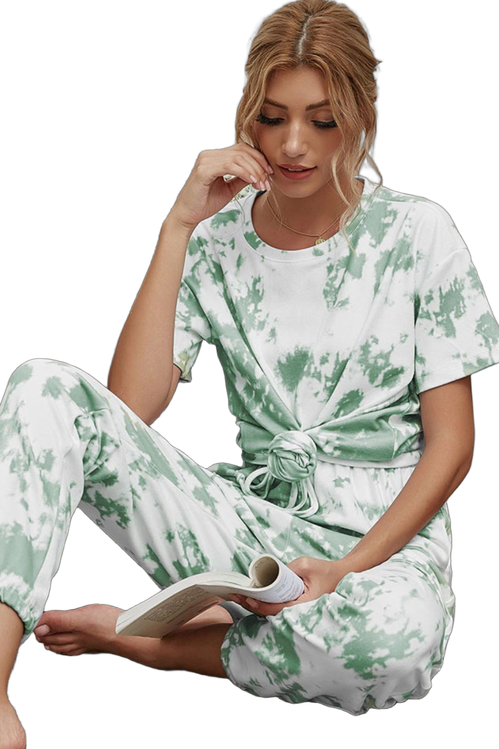 Woman wearing white and green tie-dye short-sleeve tee knotted at waist with matching drawstring joggers, seated holding a…