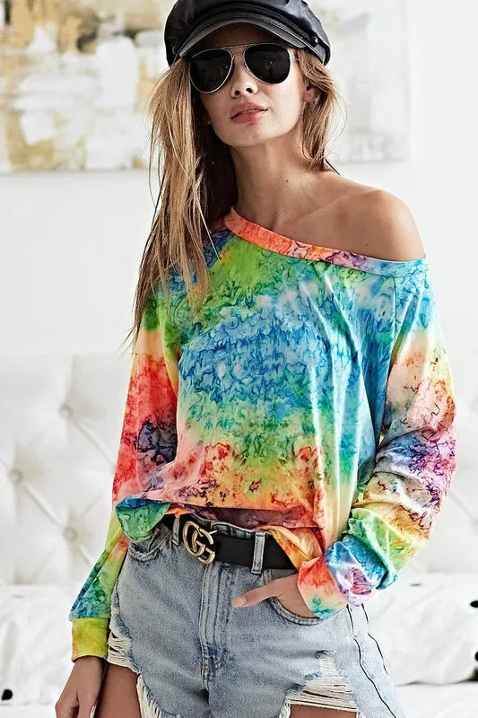 Woman wearing multicolor tie-dye long-sleeve off-shoulder top with ripped denim shorts and black belt with gold buckle
