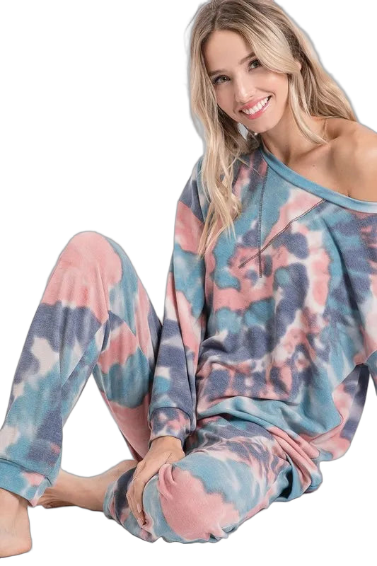 Smiling seated model wearing matching tie-dye lounge set in pink, teal and gray; off-the-shoulder top and relaxed pants