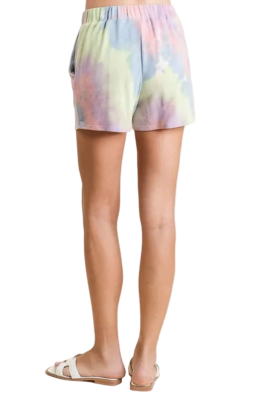 Back view of model wearing pastel tie-dye shorts with elastic waistband, side pockets, short hem, and white slide sandals