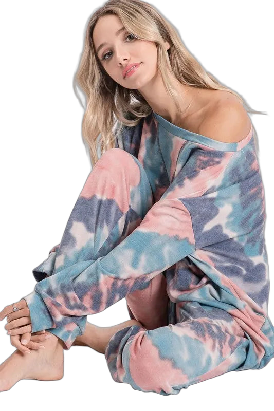 Woman seated wearing pastel pink, blue, and gray tie-dye lounge pants with matching off-shoulder top, cuffed ankles, barefoot